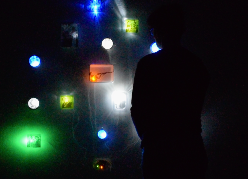 Instalacion_luminica_archivo_politica - BRISA MP Silhouetted person standing in a dark room, facing a wall of glowing colored tiles and framed images. A visitor observes a wall installation lit by blue, green, and white lights in a dark gallery.